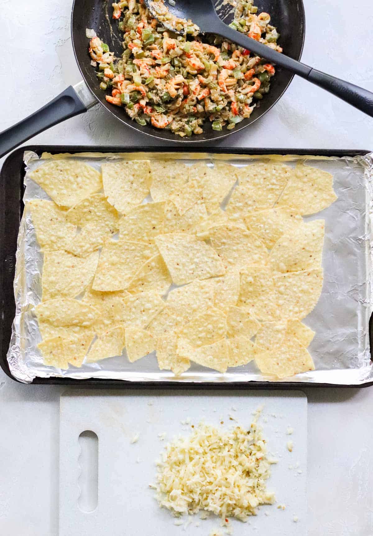 cutting board with shredded cheese, baking sheet lined with foil with tortilla chips, and a skillet full of cooked crawfish and veggies.