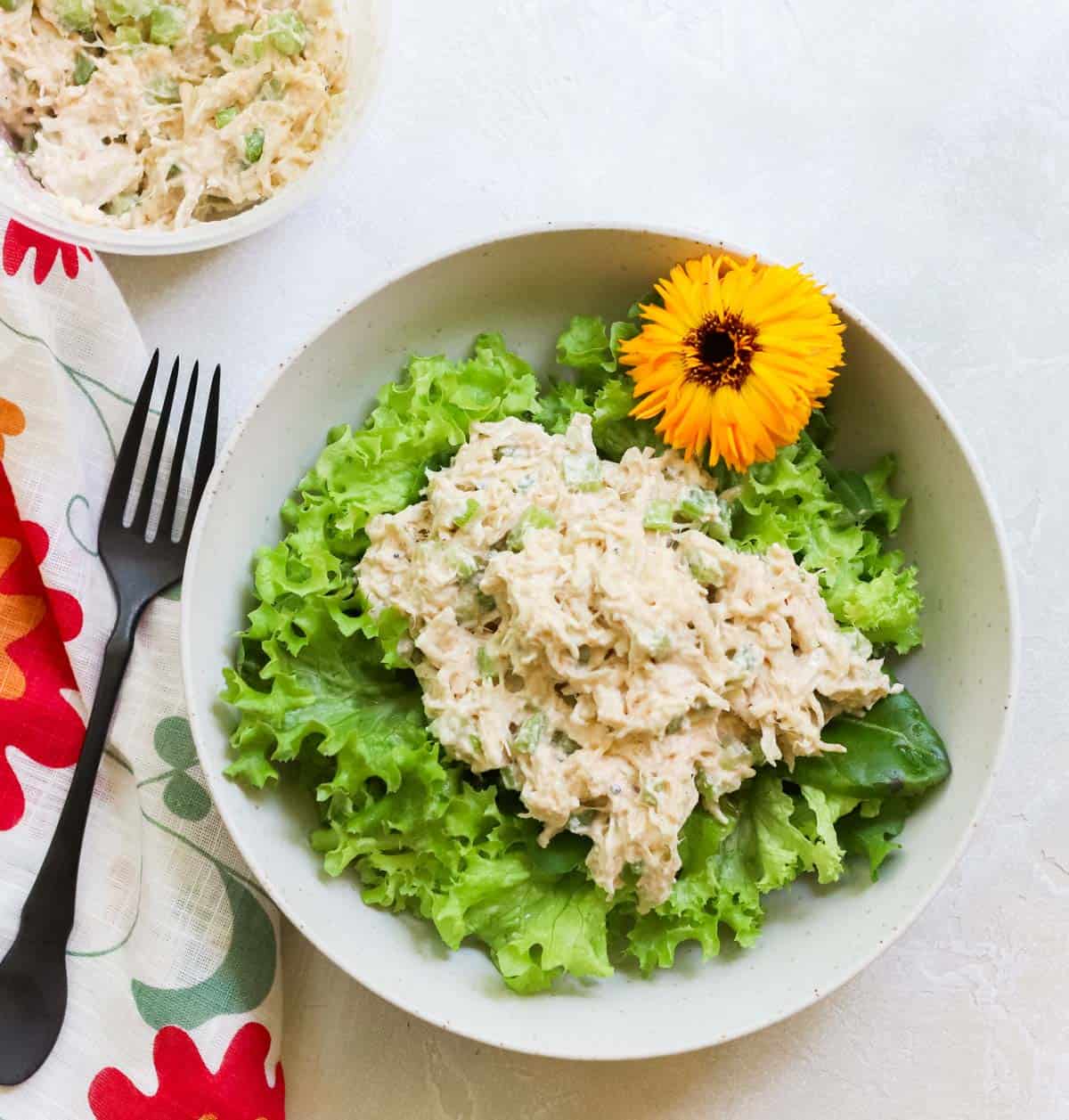bowl of lettuce with a scoop of chicken salad with an edible flower next to a black fork and a container of extra no mayo chicken salad.