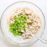 clear bowl with diced celery being stirred into chicken salad with a fork.