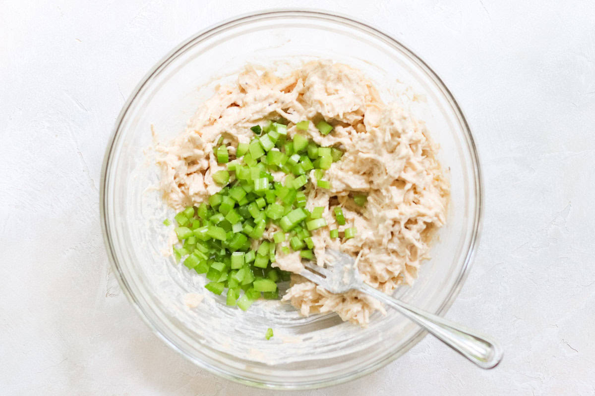 clear bowl with diced celery being stirred into chicken salad with a fork.