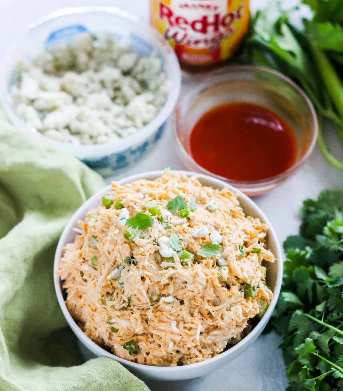 white bowl with buffalo chicken salad surrounded by a green napkin, fresh cilantro, bowl of buffalo sauce, bottle of sauce, and container of blue cheese crumbles.