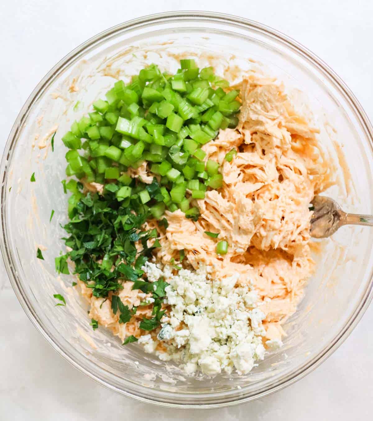 clear bowl with blue cheese crumbles, chopped celery, fresh chopped parsley, and chicken covered in yogurt and buffalo wing sauce.