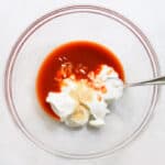 clear bowl with yogurt, buffalo wing sauce, and garlic powder with a silver spoon.