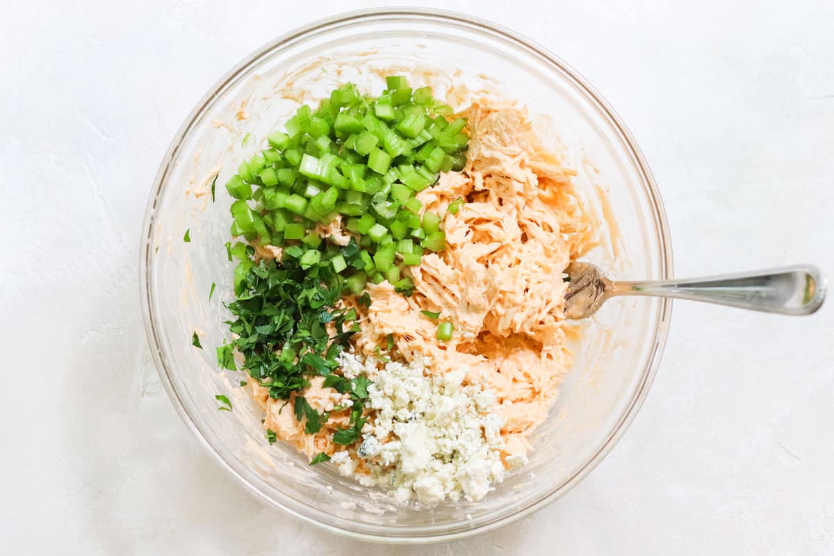 clear bowl with shredded chicken coated in buffalo wing sauce and yogurt with chopped celery, blue cheese crumbled, and fresh parsley.