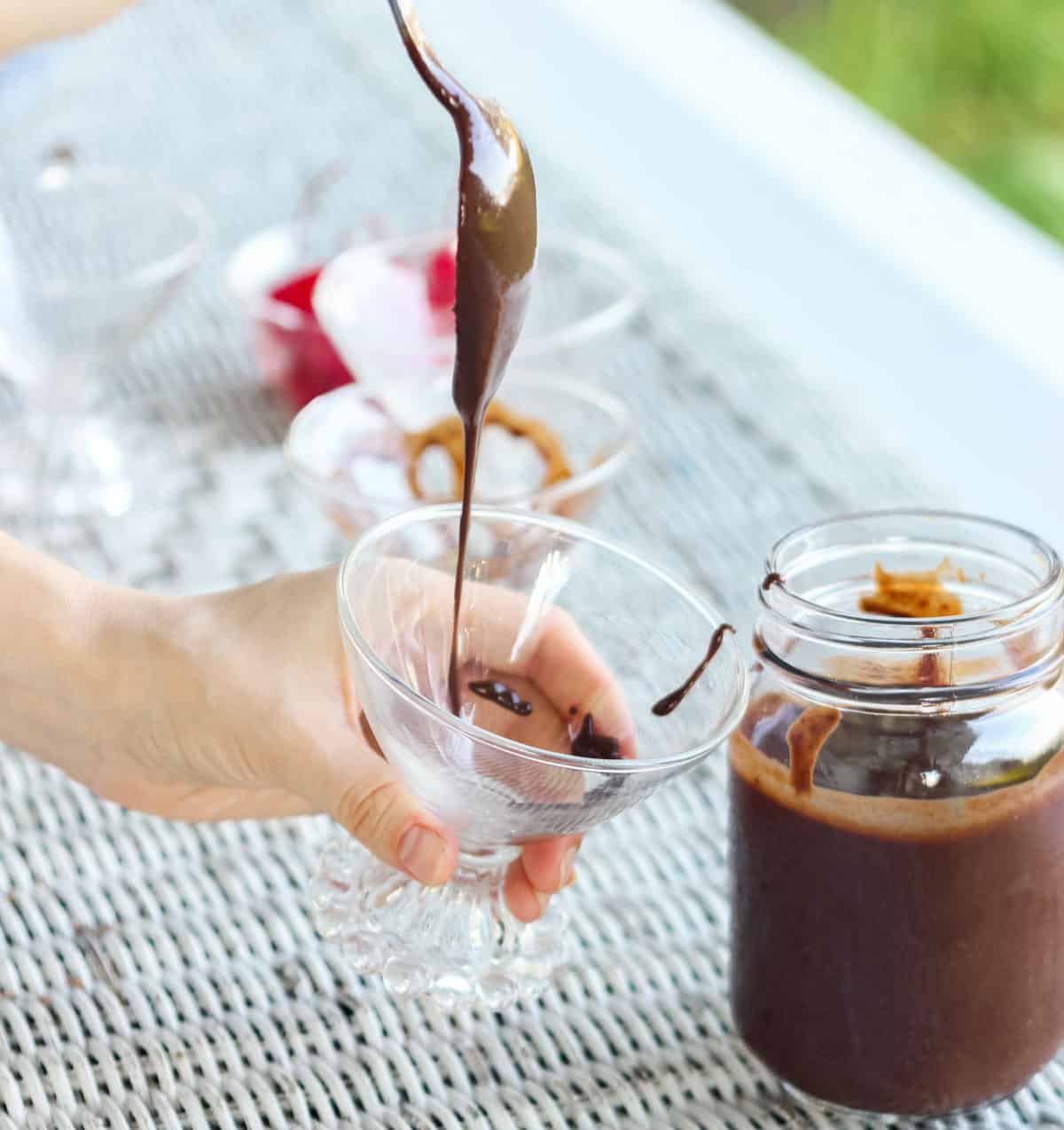 spoon drizzling chocolate sauce into a small glass next to a jar of chocolate sauce on a white wicker table.
