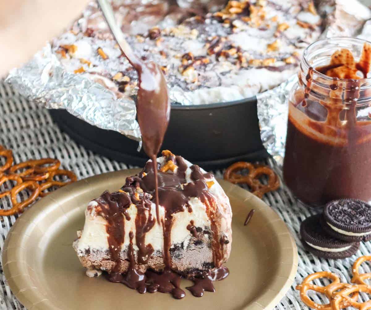gold plate with ice cream pie with a spoon drizzling chocolate sauce on top with pretzels, oreos, extra chocolate sauce, and ice cream pie in the background.