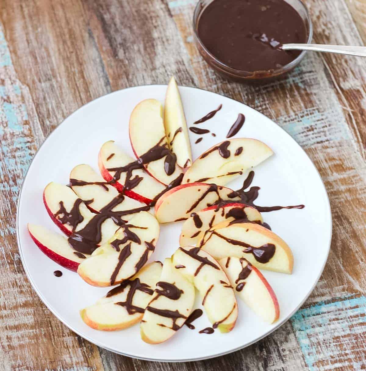wooden table with a white plate with apple slices drizzled in chocolate sauce and a small bowl of easy chocolate sauce.