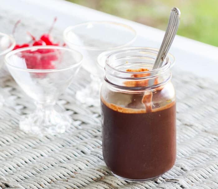 jar of chocolate sauce with a silver spoon in it on a white wicker table with drink glasses in background.