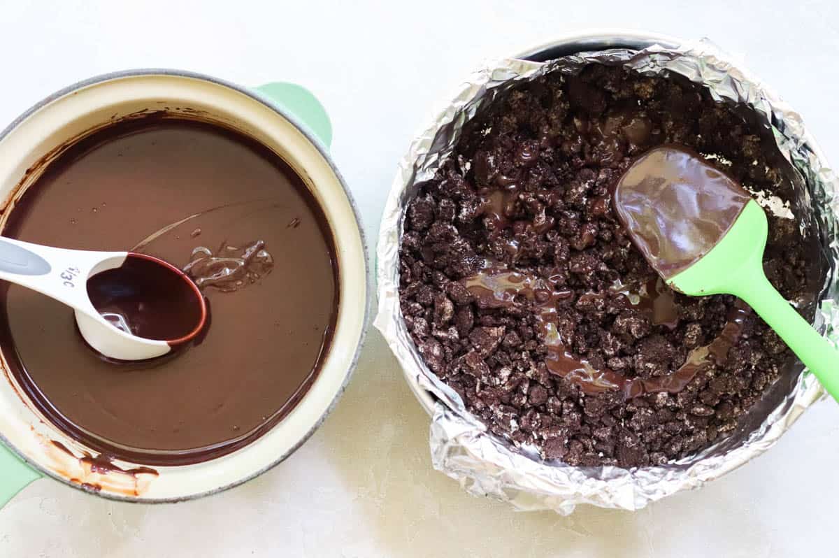 saucepan with homemade chocolate sauce next to a springform pan with crushed chocolate cookies and chocolate sauce.