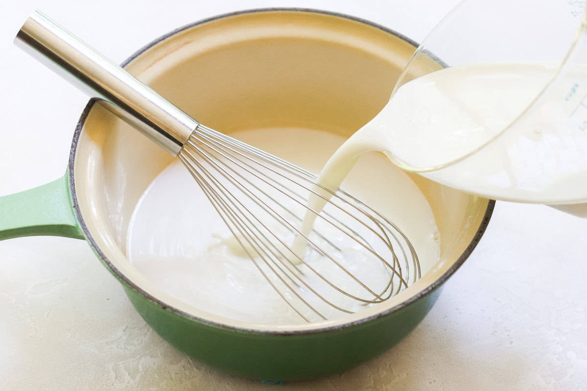 milk pouring into a green saucepan with sugar and water with a silver whisk.