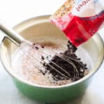 bag of chocolate chips pouring into a green saucepan with milk and sugar syrup combined with a whisk.
