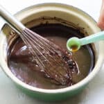 hand pouring a teaspoon of vanilla extract into chocolate sauce with a silver whisk in a green saucepan.
