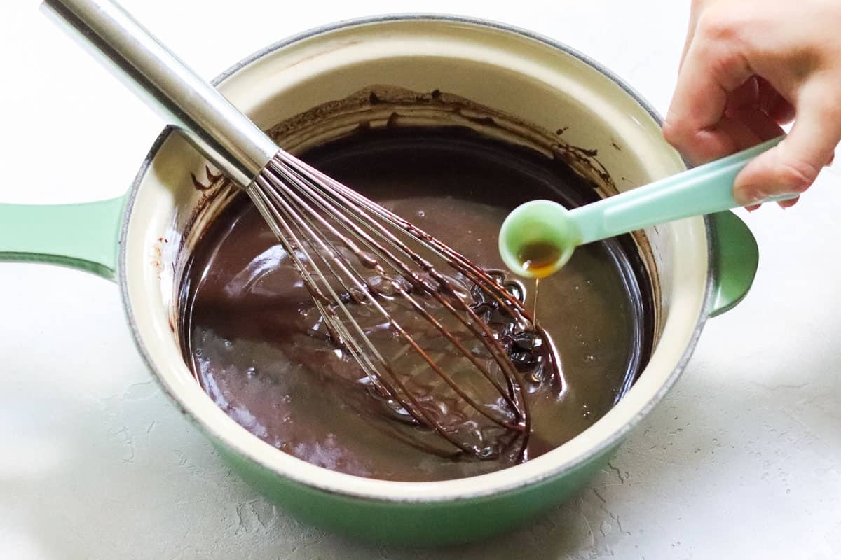 hand pouring a teaspoon of vanilla extract into chocolate sauce with a silver whisk in a green saucepan.