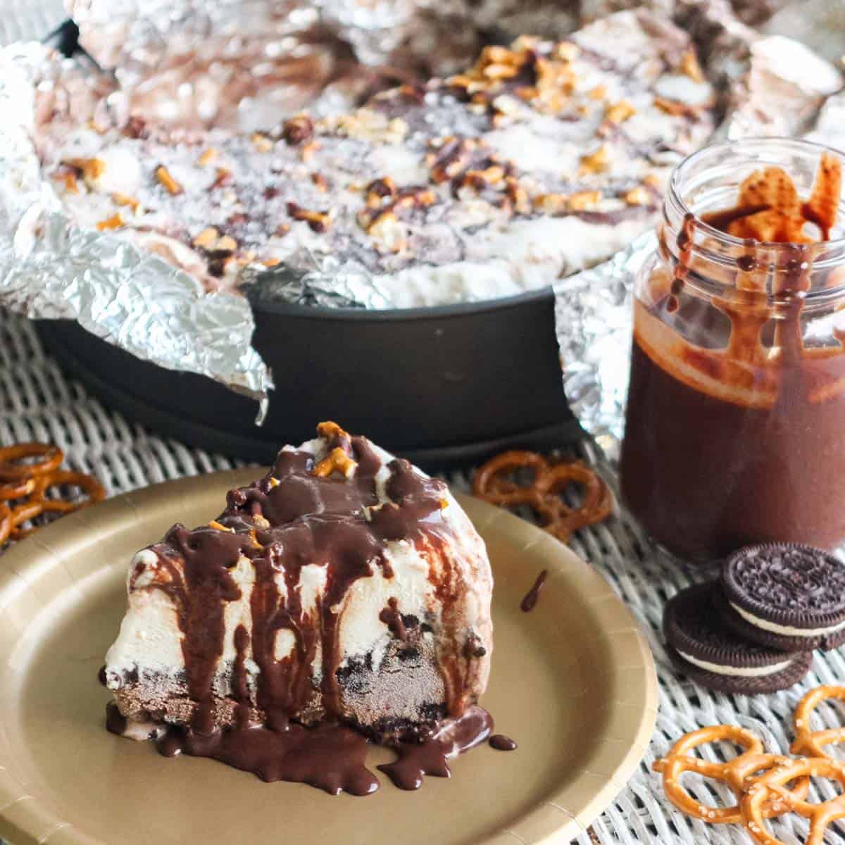gold plate with ice cream pie drizzled in chocolate sauce next to pretzels, oreos, a jar of chocolate sauce made with chocolate chips, and a pan with ice cream pie.