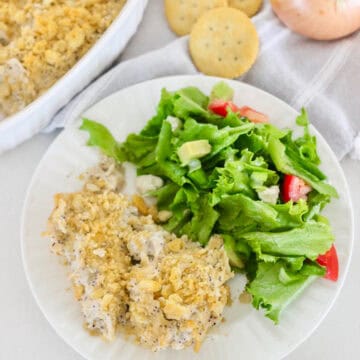 plate with poppyseed chicken casserole next to a green salad with casserole dish, ritz crackers, and an onion in background.