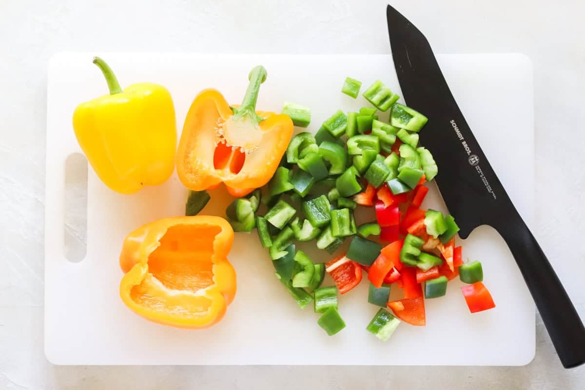 yellow, orange, green, and red bell peppers being chopped on a plastic chopping board with a black knife.