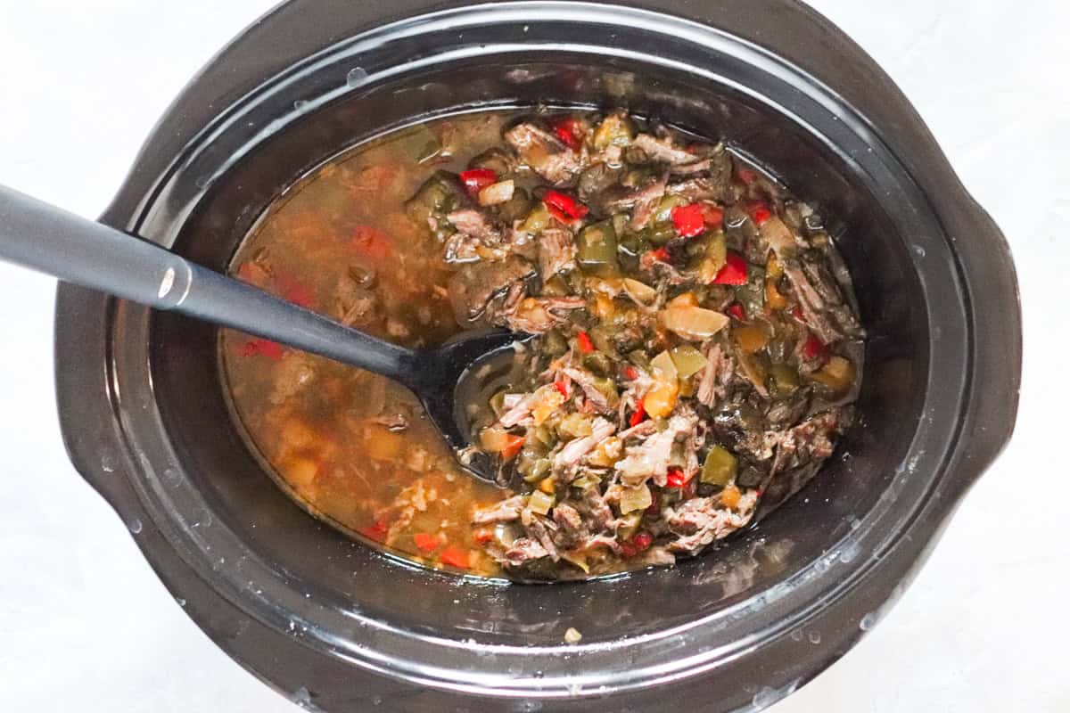 black spoon stirring together beef carnitas tacos meat in a slow cooker.