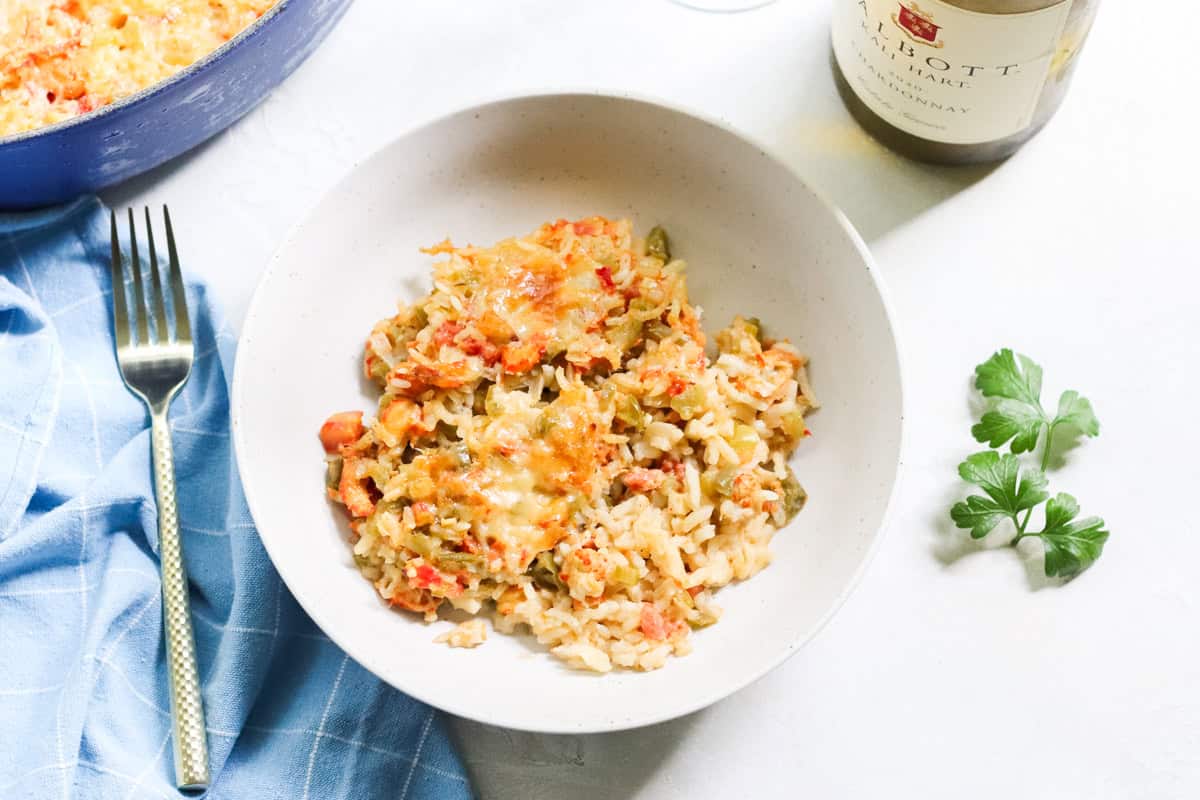 blue napkin, gold fork, blue pot, bowl with crawfish casserole, fresh parsley, empty wine glass, and bottle of chardonnay.