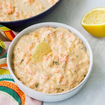 small bowl of crawfish dip with a tortilla chip next to a lemon slice, napkin, and pan of extra dip.