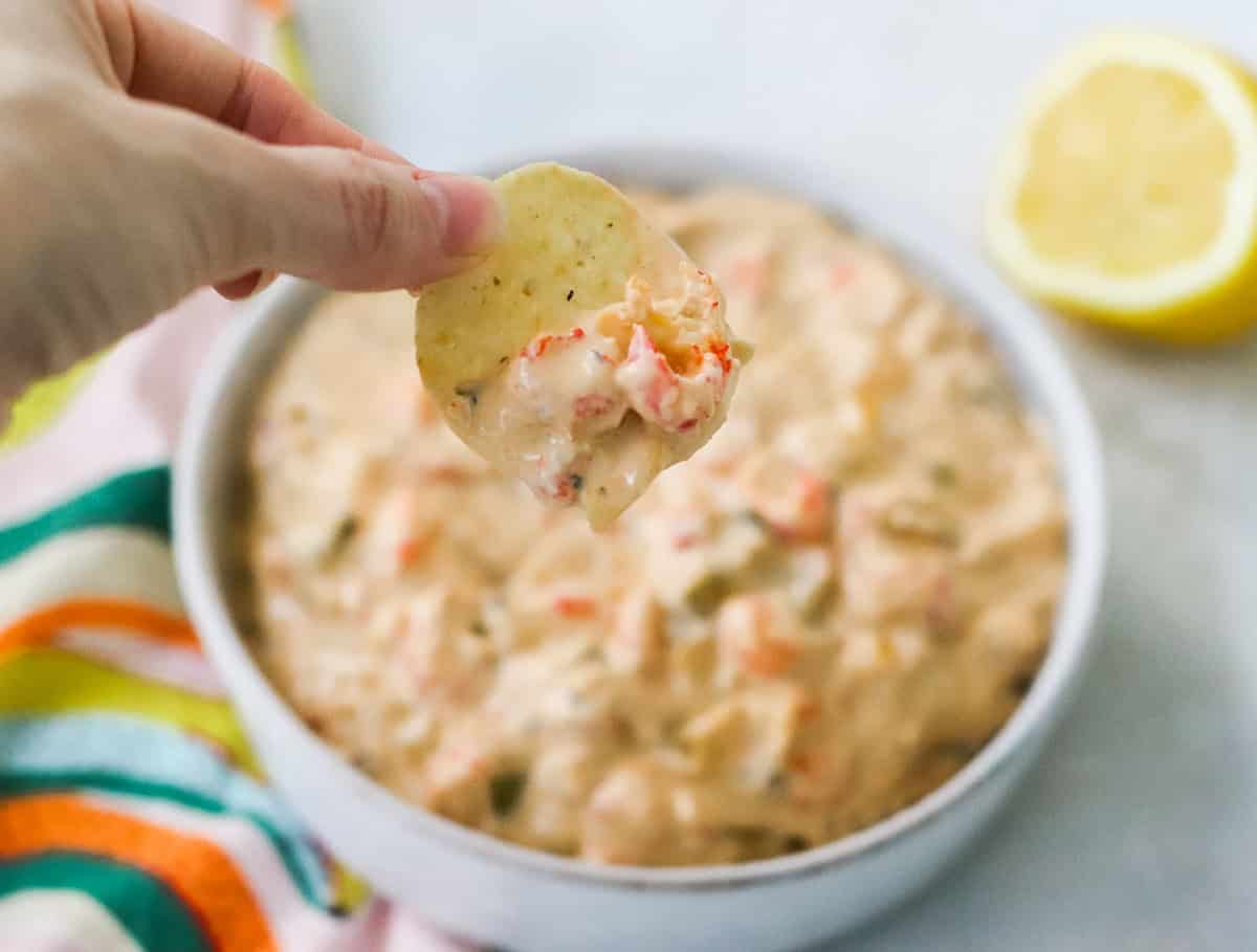 hand holding a tortilla chip with crawfish dip with a bowl of dip in background.