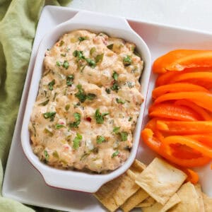 baking dish with crawfish dip on a plate with sliced orange bell peppers and pita chips next to a green napkin.
