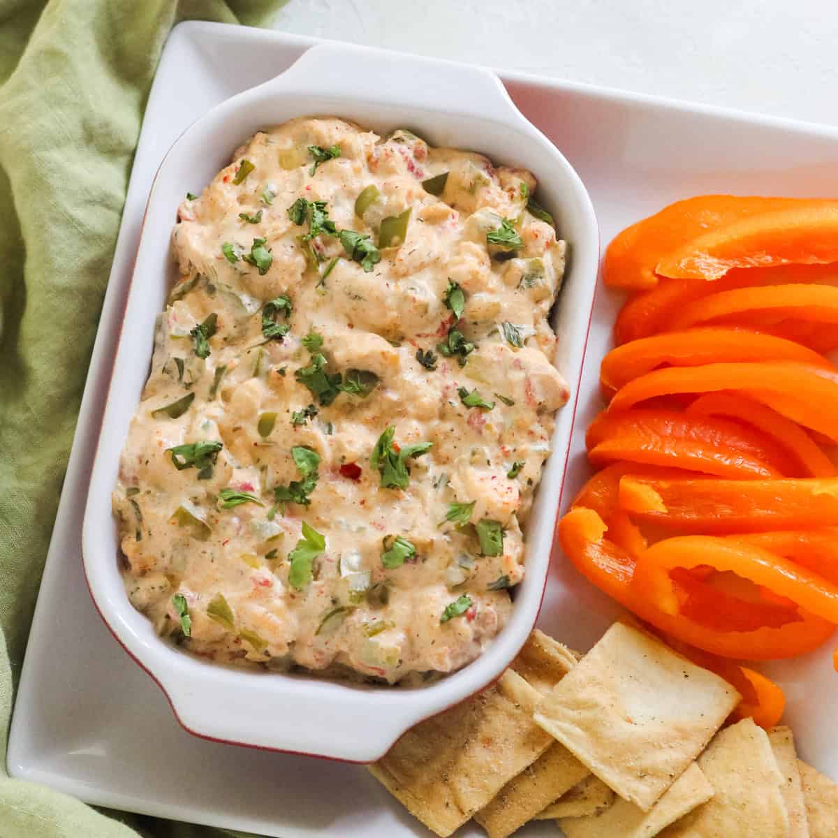 baking dish with crawfish dip on a plate with sliced orange bell peppers and pita chips next to a green napkin.