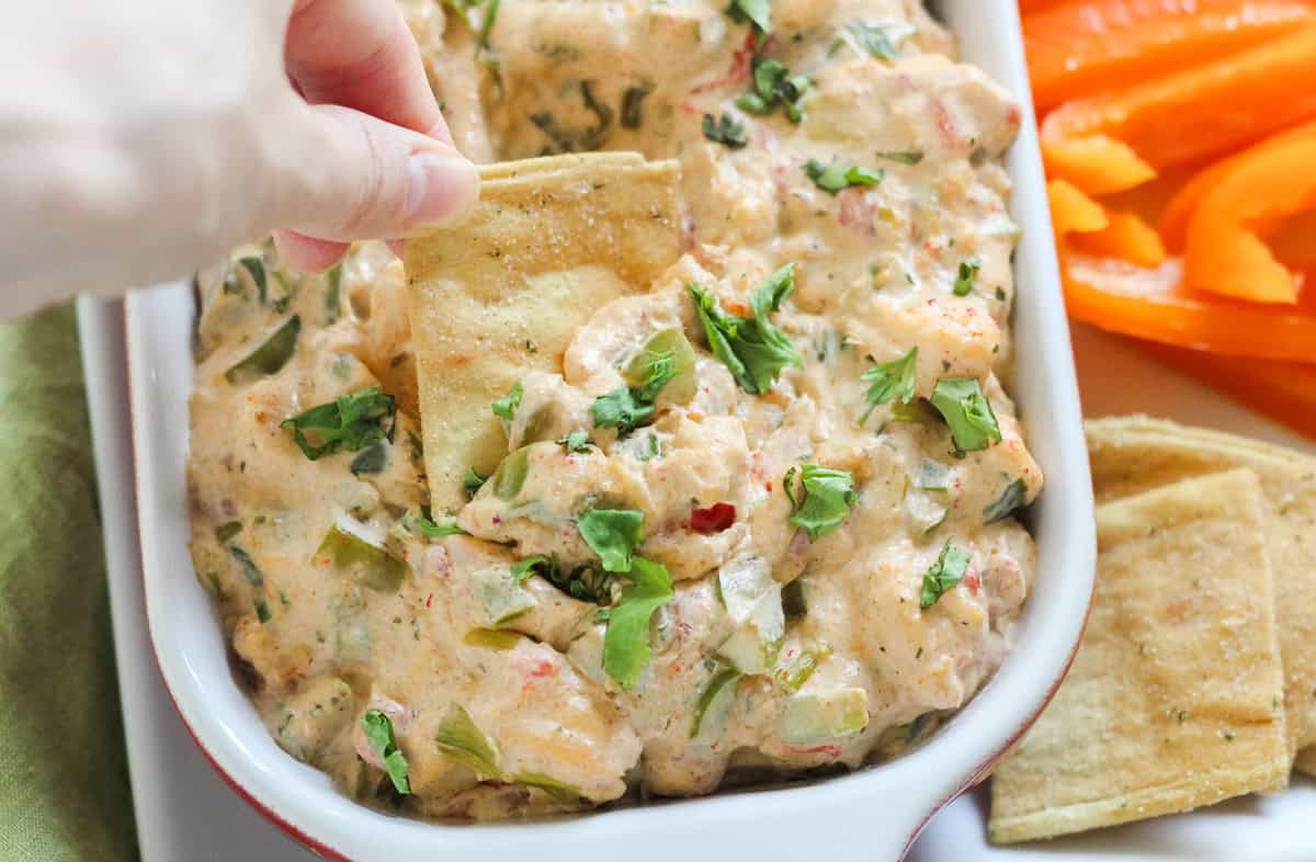 hand dipping a pita chip into a baking dish of hot crawfish dip next to extra pita chips and bell pepper slices.