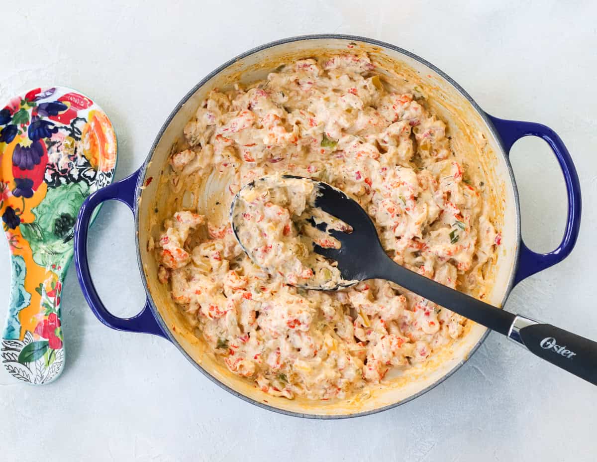 blue pan with crawfish dip being stirred by a slotted spoon next to a colorful spoon rest.