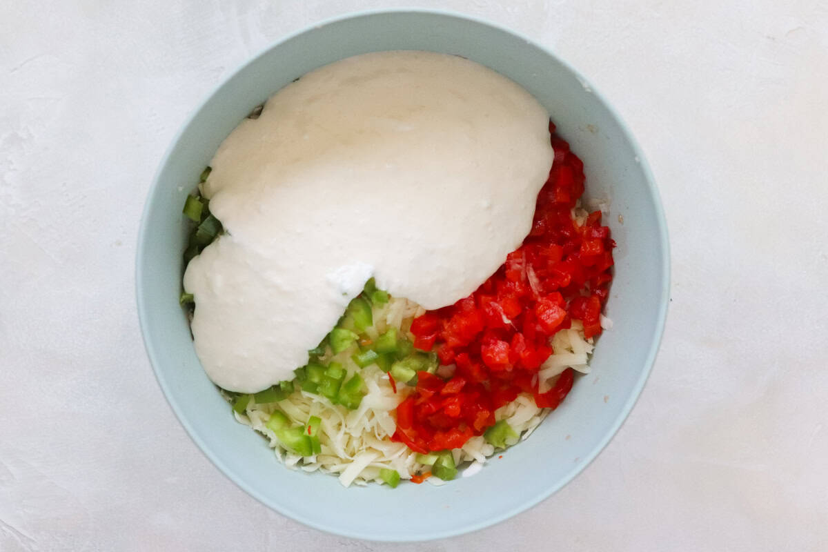 blue mixing bowl with shredded cheddar cheese, diced pimientos, minced jalapeno peppers, and blended cottage cheese.