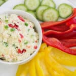 bowl of jalapeno pimento cheese next to yellow and red bell peppers plus cucumber slices.