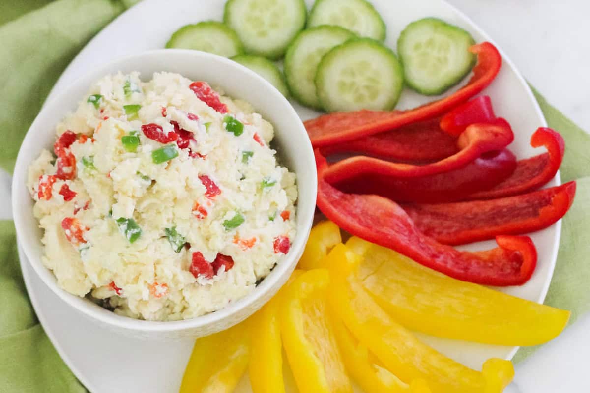 bowl of jalapeno pimento cheese next to yellow and red bell peppers plus cucumber slices.