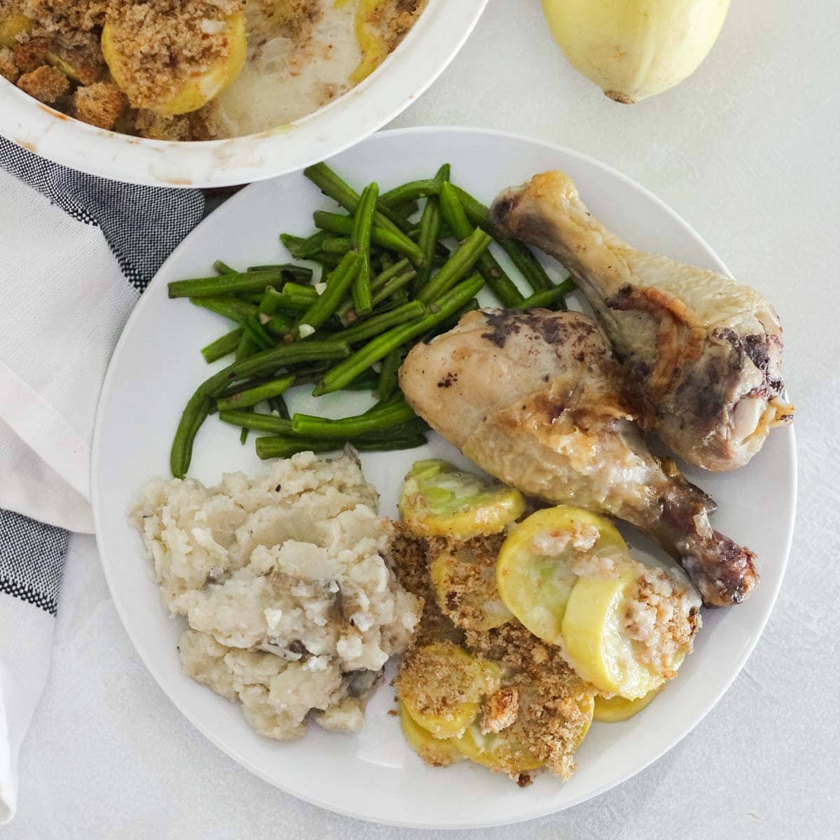 plate with mashed potatoes, squash casserole, sauteed green beans, and fried chicken with a casserole of squash casserole in background.