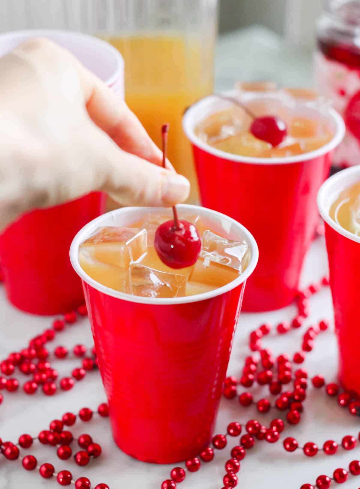 hand placing cherry into yellow hammer drink with a pitcher of cocktails in background.