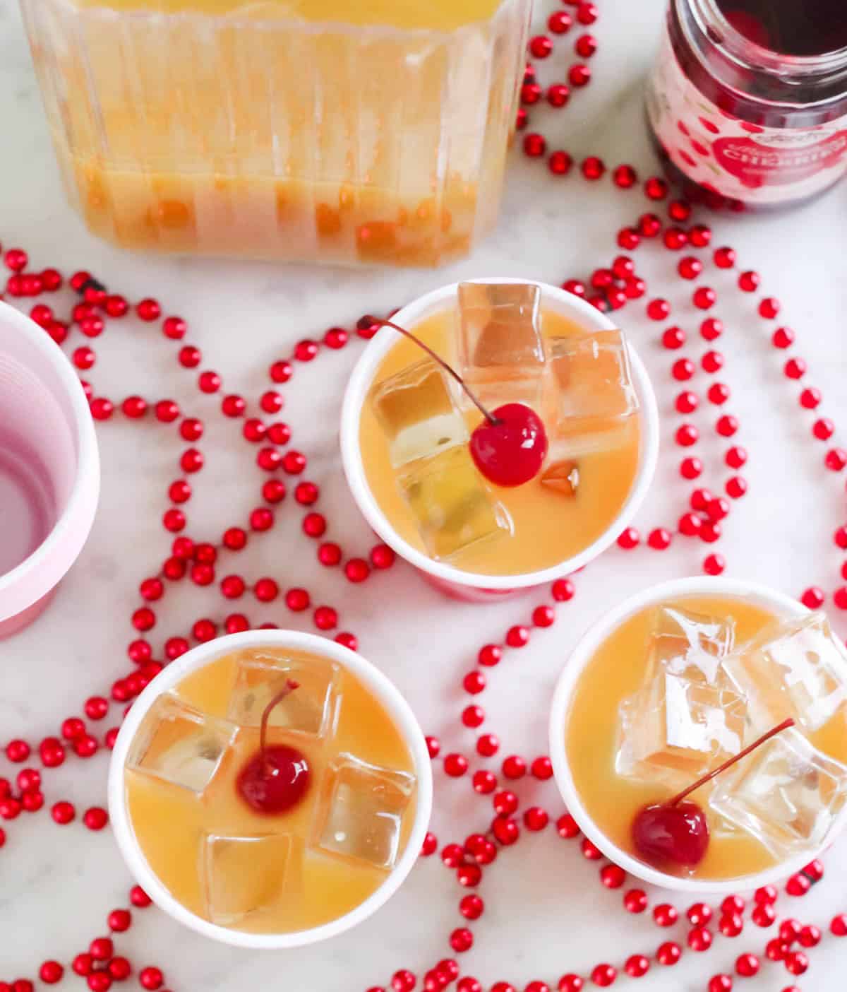overhead view of three yellow hammer drinks with cherries, red beads, pitcher and cherries.