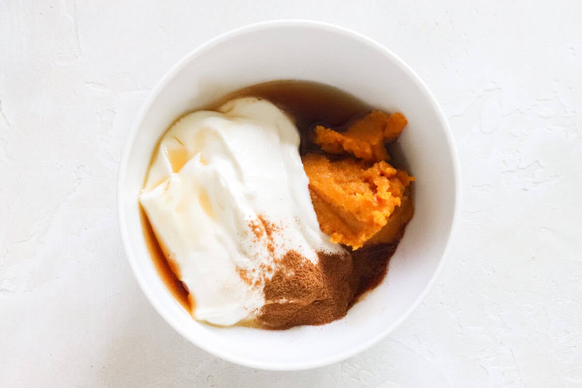 white bowl with greek yogurt, cinnamon, pumpkin puree, maple syrup on white counter.