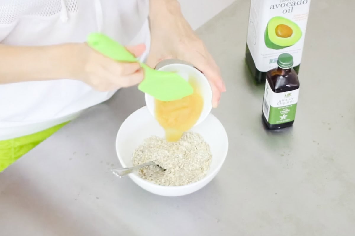 hand pushing apple sauce into a bowl of microwave oatmeal cookie dry ingredients with a green spatula.