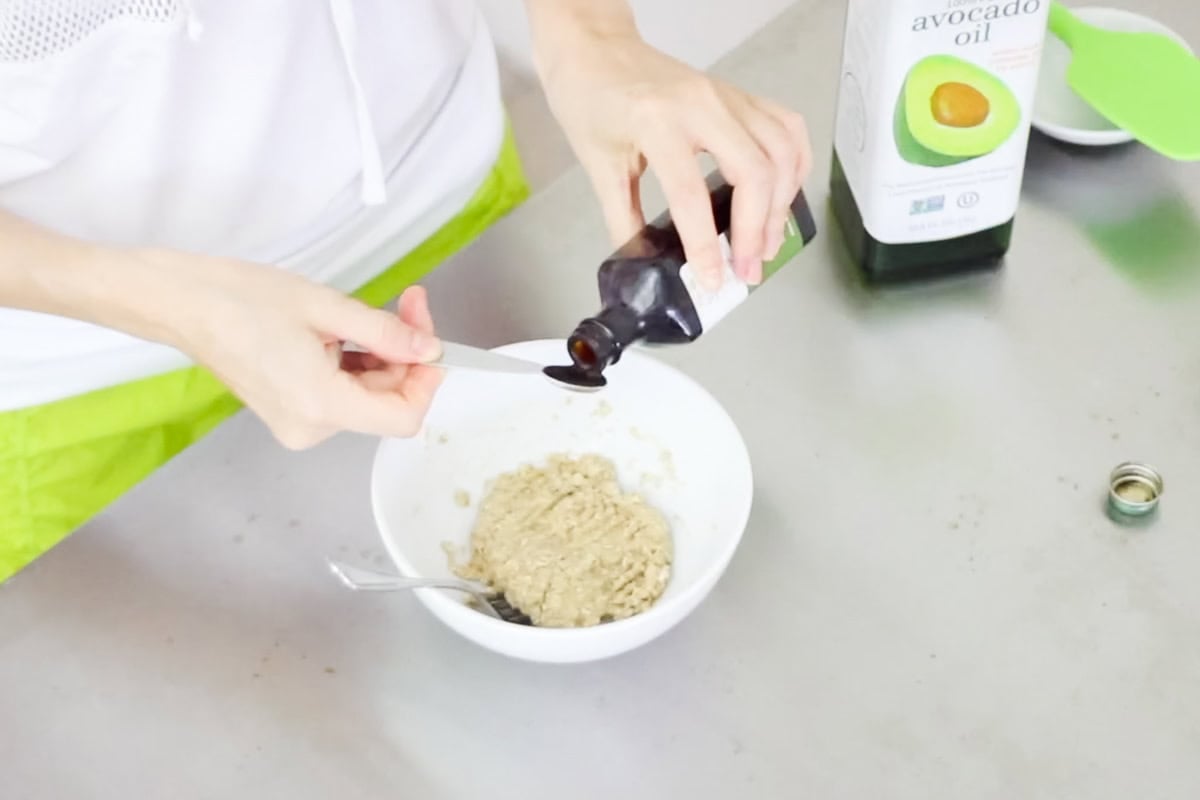 hand pouring vanilla extract into a measuring spoon over a bowl of cookie dough in a mug.