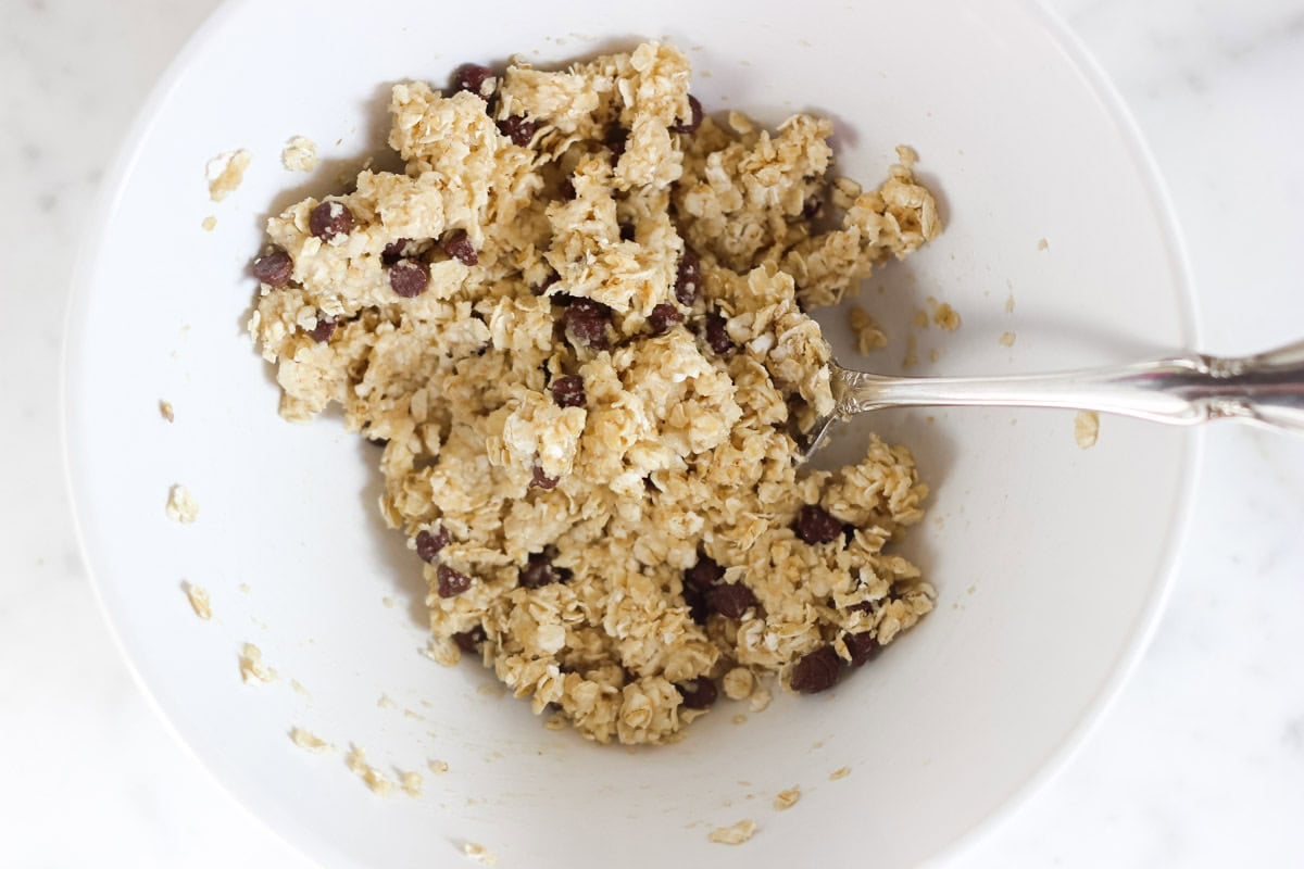 white bowl with oatmeal cookie dough ingredients being stirred together with a silver spoon.